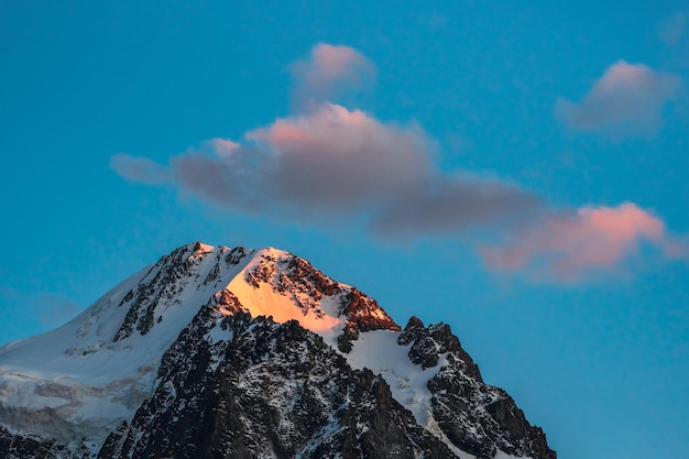 Top black rocks in evening golden sunshine and white-snow pointy peak. Atmospheric dawn landscape with high snowy mountain with peaked top.