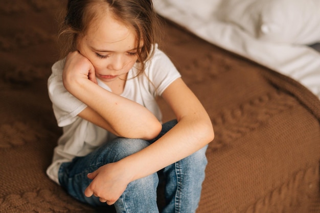 Top closeup view of distraught beautiful little girl with wet eyes from tears sits on bed in bedroom and looking down