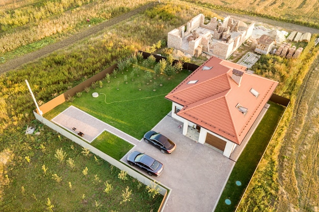 Top down aerial view of two private houses, one under construction with concrete foundament and brick walls and another finished with red tiled roof.