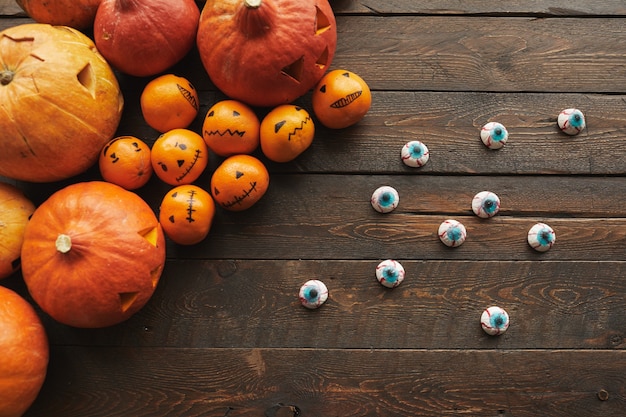 Top-down flat lay composition of carved pumpkins, tangerines with jack o' lantern faces and scary eyeball candies lying on wooden table