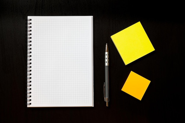 Top down flat lay view of notepad with white pages pen and sticky note blocks on black background