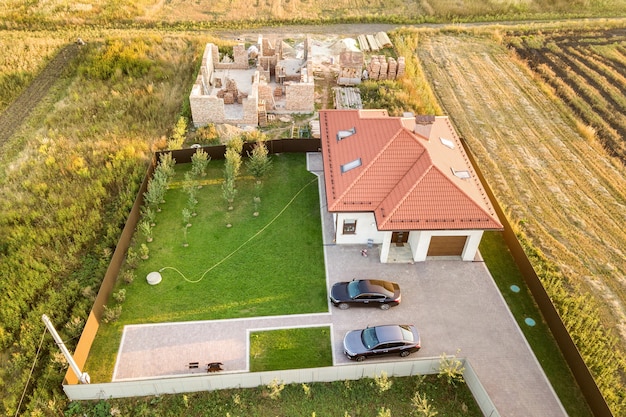 Top-down luchtfoto van twee particuliere huizen, een in aanbouw met betonnen fundering en bakstenen muren en een andere afgewerkt met een rood pannendak.