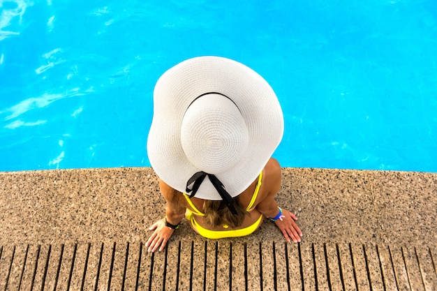 Top down mening van jonge vrouw die gele strohoed dragen die dichtbij zwembad met duidelijk blauw water rusten op de zomer zonnige dag.