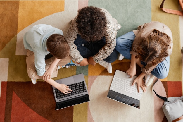 Вид сверху на группу из трех детей, использующих компьютеры, сидя на ковре на полу