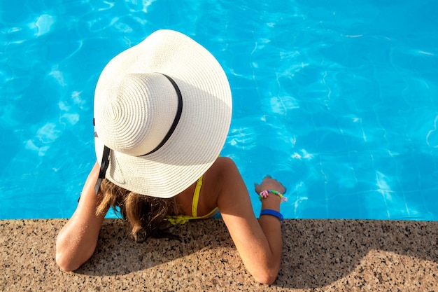 Top down view of young woman wearing yellow straw hat resting in swimming pool with clear blue water on summer sunny day.
