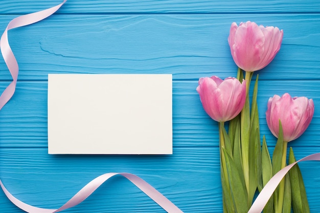Top flat lay overhead view photo of pink tender tulips with ribbon and empty paper white greeting card for text on bright blue desk