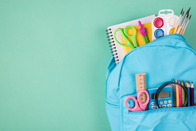 Top above overhead view photo of blue backpack filled with colorful stationery placed to the right side isolated on turquoise background with copyspace