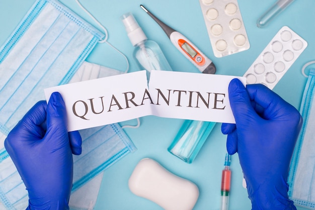 Top above overhead view photo of doc hands in gloves tearing quarantine word with masks sanitizer pills soap and shots isolated on blue background