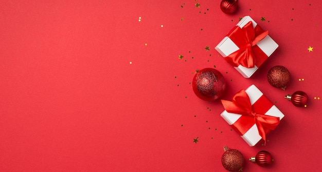 Top panoramisch uitzicht foto van witte geschenkdozen met rode satijnen lint bogen kerstboom ballen en gouden confetti op geïsoleerde rode achtergrond met copyspace