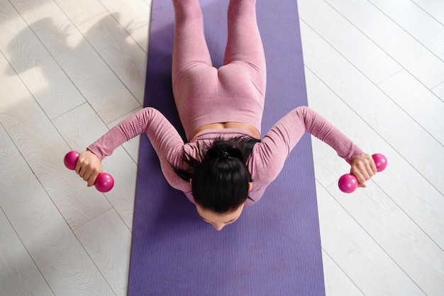 Вид сверху на активную молодую женщину в стильной спортивной одежде, тренирующуюся с гантелями, лежащую на коврике для йоги дома Концепция людей и здорового образа жизни