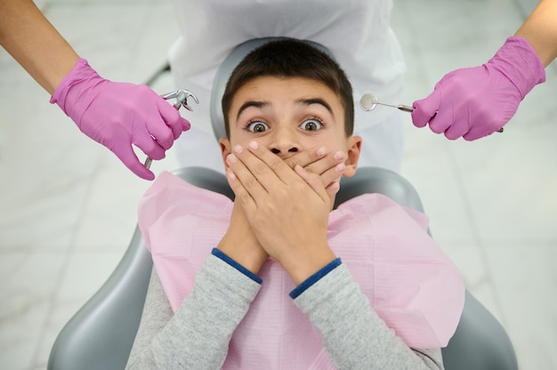 Вид сверху испуганного мальчика, прикрывающего рот от страха руками, сидя на кресле стоматолога на фоне рук в розовых хирургических перчатках, держа стоматологические инструменты из нержавеющей стали возле его лица