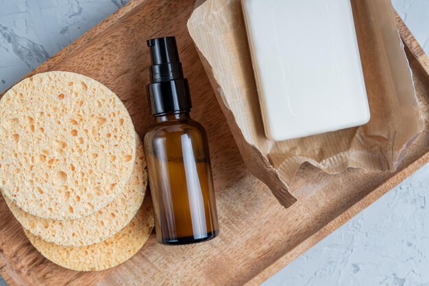 Top view amber glass cream bottle, homemade soap, face sponges on wooden board. SPA natural organic beauty product packaging design.