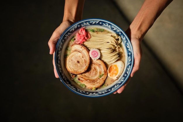 Top view of anonymous woman showing bowl of delicious ramen garnished with pork and egg against dark blurred background