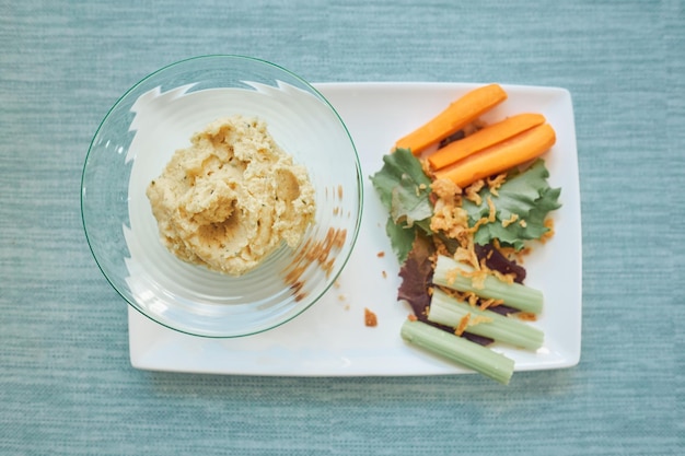 Top view of appetizing hummus in bowl served on white plate with celery lettuce and carrot on table with blue tablecloth