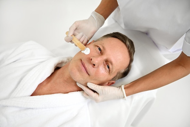 Top view of attractive handsome mature man, relaxing on a massage table at wellness spa salon and looking at camera while beautician applying nourishing cosmetic product on his face. Male beauty care