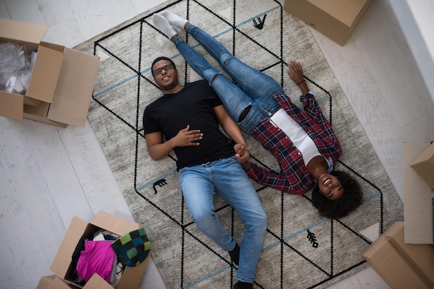 Вид сверху на привлекательную молодую афроамериканскую пару, которая движется, держится за руки, смотрит в камеру и улыбается, лежа среди картонных коробок