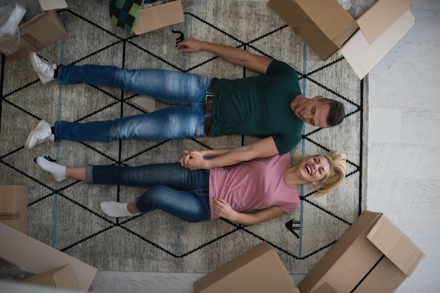 Photo top view of attractive young couple moving, holding hands, looking at camera and smiling while lying among cardboard boxes