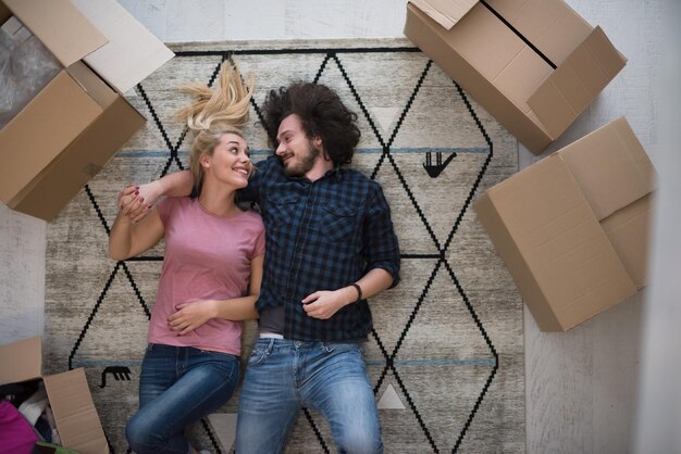 Top view of attractive young couple moving, holding hands, looking at camera and smiling while lying among cardboard boxes