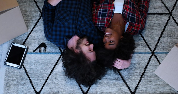 Top view of attractive young multiethnic couple moving in, holding hands, looking at camera and smiling while lying among cardboard boxes
