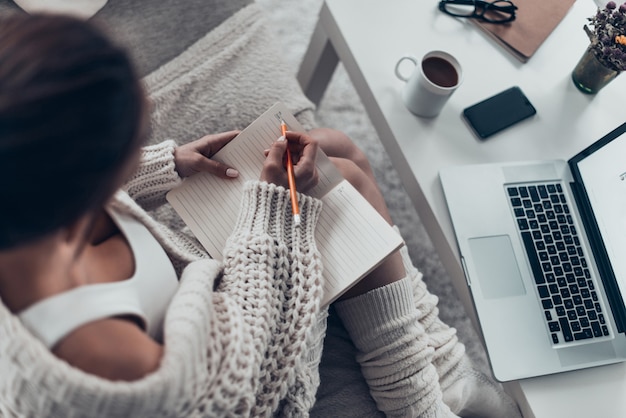 Top view of attractive young woman writing something in her note pad while spending weekend at home