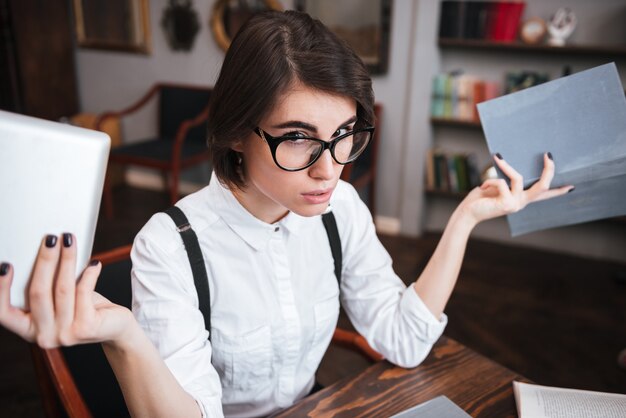 Top view of Authoress in glasses and white shirt sitting by the table and choosing between books