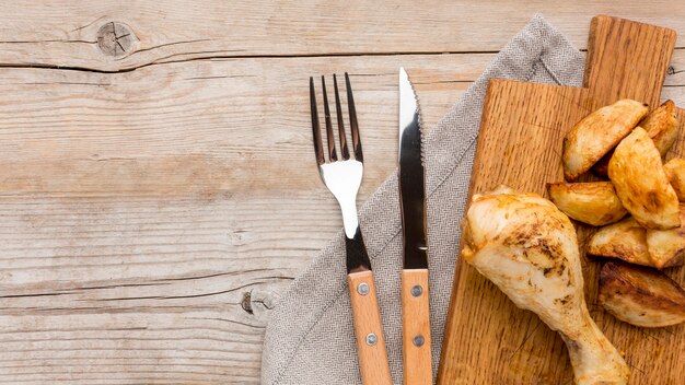 Top view baked chicken drumstick and potatoes on cutting board with cutlery and copy-space