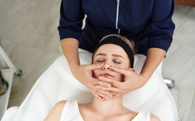 Top view of beautician doing facial anti-aging lifting massage on woman face at spa clinic. Professional lymphatic drainage massage at modern spa center