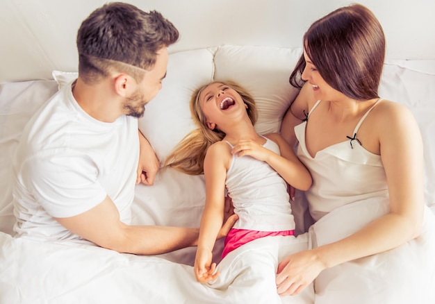 Top view of beautiful family in bed in the morning. Little daughter is laughing while her parents are tickling her