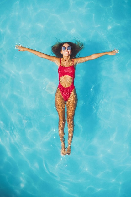 Top view of a beautiful smiling happy young woman enjoying in the water of the pool. She is relaxing and swimming in clear blue water and smiling.