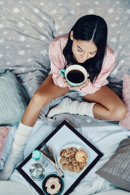 Top view of beautiful young woman in pajamas enjoying hot drink while resting in bed at home