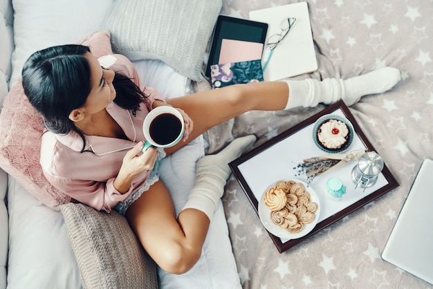 Top view of beautiful young woman in pajamas enjoying hot drink while resting in bed at home