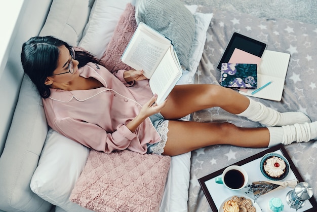 Top view of beautiful young woman in pajamas reading book while resting in bed at home