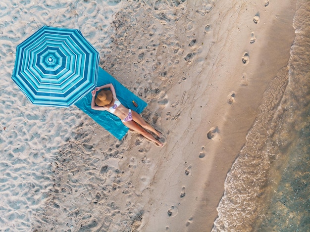 Top view of a beautiful young woman who is enjoying in sunbathe on the beach dressed in a bikini. She is relaxing on the blue beach towel under the blue umbrella.