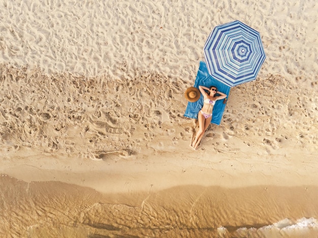 海のビーチで日光浴を楽しんでいる美しい若い女性の平面図です。彼女は青い傘の下で青いビーチタオルの上でリラックスしています。