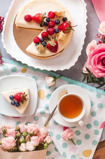 Top view of a berry cheesecake with a cup of tea surrounded by fresh flowers