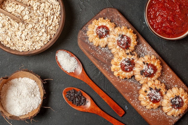 top view biscuits with jam on wood board coconut powder and chocolate powder in wooden spoons flour oats and jam in wooden bowls on dark background