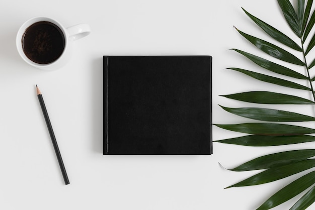 Photo top view of a black book mockup with workspace accessories and a palm leaf on a white table