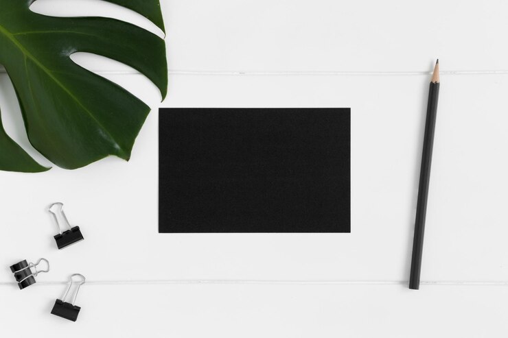 Photo top view of a black card mockup with workspace accessories and a monstera leaf on a white table