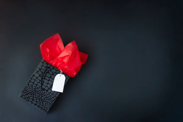 Top view of black gift bag with red tissue paper and white label on black background