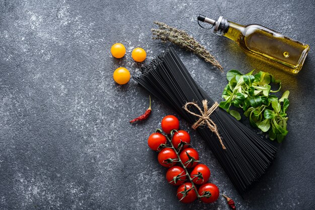 Top view of black pasta, tomatoes, olive oil, lettuce, chili peppers on grey background with copy space, cooking dinner concept