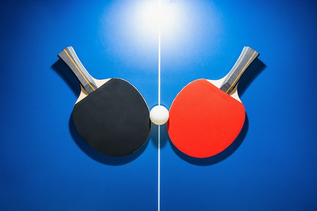 Top view black and red table tennis racket and a white ping pong ball on the blue ping pong table with a bright spotlight, Two table tennis paddle is a sports competition equipment for indoor exercise