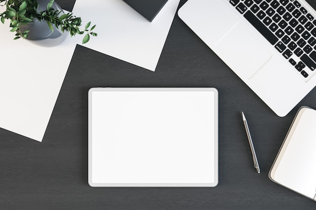 Top view on blank white digital tablet screen with on black wooden table surface with laptop green plant and paper sheets 3D rendering mock up