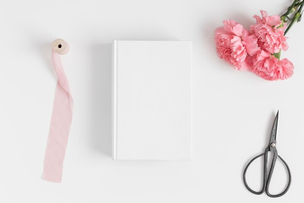 Photo top view of a book mockup with a bouquet of pink carnations and workspace accessories on a white table