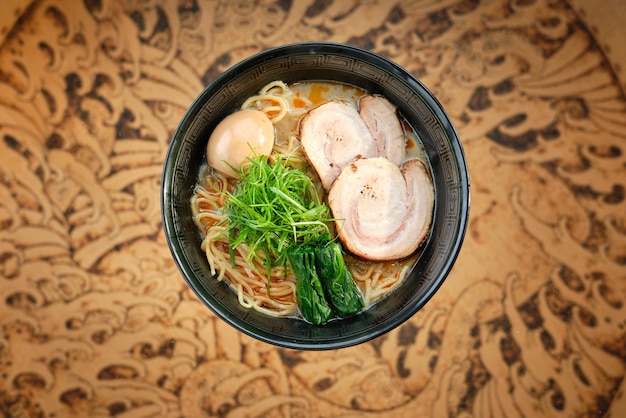 Top view of bowl of appetizing Japanese ramen with noodles and broth garnished with rolled pork nori and egg and served on carved wooden table in restaurant