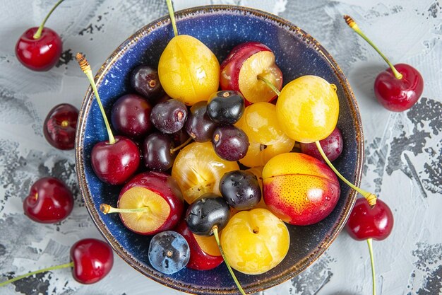 Top view of bowl with berries fresh ripe fruits with yellow cherries and plums on grey