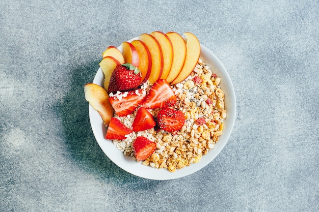 Top view of breakfast porridge oatmeal with granola flakes fruits strawberries and peaches in a white plate on a grey background