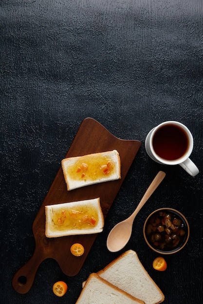 Top view of breakfast set with jam smeared on bread slices on cutting board with jam bread slices cup of tea kumquats and spoon on black background with copy space