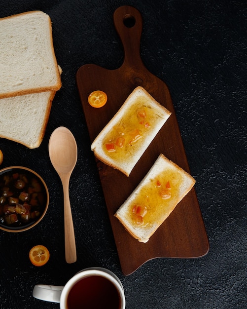 Top view of breakfast set with jam smeared on bread slices on cutting board with jam bread slices cup of tea kumquats and spoon on black background