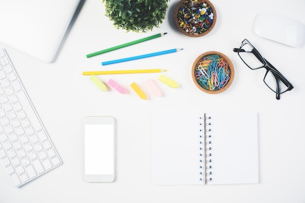 Top view of bright desktop with blank white cellphone, spiral notepad, colorful supplies, glasses keyboard and plant. Mock up