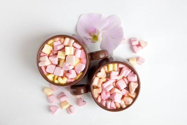 Top view of brown cups of hot cocoa with marshmallows in heart\
shape on white background romantic background with hot chocolate\
and orchid flower love concept for valentine s day or wedding